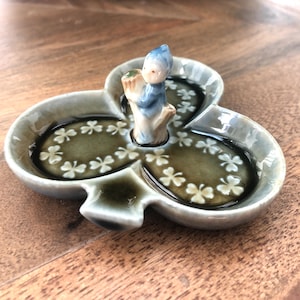 May include: A ceramic, three-section dish in a cloverleaf shape. The dish is light grey with a dark green interior decorated with white clover designs. A small figurine of a child in blue and white clothing stands in the centre.
