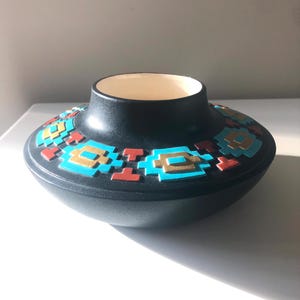 May include: A black ceramic bowl with a wide, shallow base and a smaller, cylindrical top. The bowl is decorated with a geometric pattern in turquoise, gold, and red. The interior of the bowl is white.