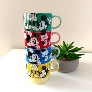 May include: A stack of four colorful ceramic mugs featuring Mickey Mouse. Each mug is a different color: green, red, blue, and yellow. The mugs have matching colored handles and are stacked on top of each other.