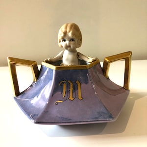 May include: A vintage porcelain doll figurine inside a lavender-coloured, hexagonal-shaped container with gold trim and handles. The container has a gold letter "M" on the front.