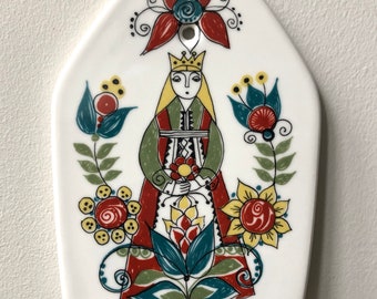May include: White ceramic decorative plaque with a folk art design. The design features a crowned figure in a red and green dress, surrounded by colorful flowers and foliage. A hole is at the top for hanging.