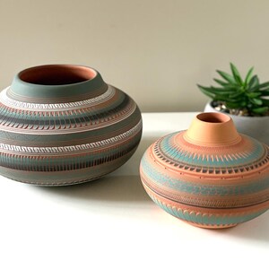 Peut inclure: Deux vases en céramique faits à la main avec des motifs géométriques. Le plus grand vase est principalement gris-vert avec des rayures marron et blanches. Le plus petit vase est pêche avec des rayures turquoise et marron. Une petite plante succulente est en arrière-plan.