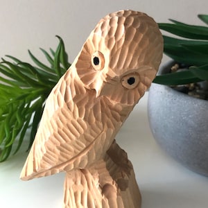 May include: A carved wooden owl figurine with a textured surface. The owl has black eyes and is perched on a small wooden base. The carving has a natural wood tone and is set against a neutral background.