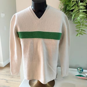 May include: Cream V-neck sweater with a green stripe across the chest. The sweater is displayed on a black mannequin. The ribbed knit fabric is visible. The sweater is a classic design.