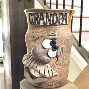 May include: A brown ceramic vase with the word "GRANDPA" in raised letters. The vase features a whimsical face design with black eyes, a large nose, and a smiling mouth. The vase is on a wooden surface.