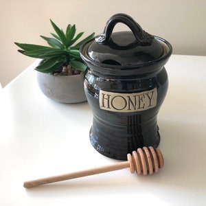 May include: A black ceramic honey pot with a lid and handle, labelled "HONEY" on a plaque. A wooden honey dipper is in front. A small potted plant is in the background.