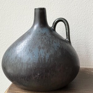 Peut inclure: Un vase en céramique sombre et bulbeux avec une anse. Le vase présente une finition texturée et mouchetée dans des tons de noir et de bleu. Le vase est posé sur une surface en bois.