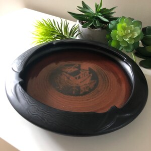 May include: A black ceramic ashtray with a brown interior. The ashtray is circular with a wavy design on the rim. The interior has a textured, concentric circle pattern.