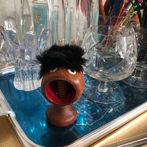 May include: A novelty nutcracker with a wooden body, red interior, and a black fur wig. The nutcracker has a screw mechanism and cartoon-like eyes. It sits on a blue tray with other glassware in the background.