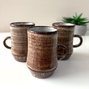 May include: Three brown ceramic mugs with handles. The mugs have a speckled, textured surface and a dark brown rim. The base of each mug features vertical lines. A small green plant is visible in the background.