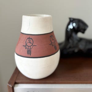 May include: A handcrafted, cream and terracotta ceramic vase. The vase features a band of terracotta with black dreamcatcher designs. The vase has a rounded base and a slightly flared top. A black panther statue is in the background.