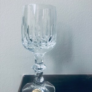 Puede incluir: Una copa de vino de cristal transparente con l&iacute;neas grabadas verticales. La copa tiene una base redonda, un tallo decorativo y una peque&ntilde;a pegatina dorada que dice "24% Bleikristall GERMANY". La copa est&aacute; sobre una superficie marr&oacute;n oscuro.