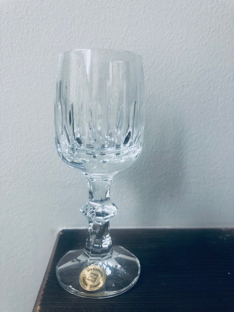 Puede incluir: Una copa de vino de cristal transparente con l&iacute;neas grabadas verticales. La copa tiene una base redonda, un tallo decorativo y una peque&ntilde;a pegatina dorada que dice "24% Bleikristall GERMANY". La copa est&aacute; sobre una superficie marr&oacute;n oscuro.