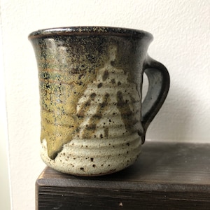 May include: Handmade ceramic mug with a dark brown rim and handle. The mug has a textured surface with brown, beige, and olive green tones. The design includes speckled details and a unique, organic shape.