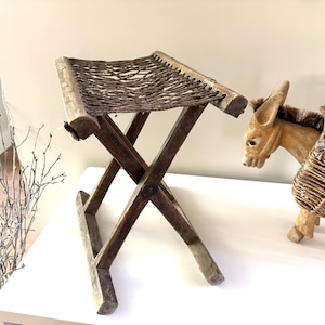May include: A folding stool with a dark brown wooden X-frame base and a woven dark brown fibre seat. Beside the stool stands a carved wooden donkey figurine featuring a dark mane and two woven wicker baskets attached to its back with rope.