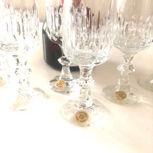 Puede incluir: Varios vasos de vino de cristal transparente con intrincados patrones de corte vertical. Las copas tienen un dise&ntilde;o cl&aacute;sico con una base redonda y un tallo decorativo. Una botella de vino parcialmente visible est&aacute; en el fondo. Cada copa tiene una peque&ntilde;a pegatina en la base.