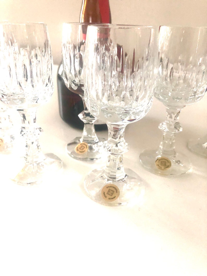 Puede incluir: Varios vasos de vino de cristal transparente con intrincados patrones de corte vertical. Las copas tienen un dise&ntilde;o cl&aacute;sico con una base redonda y un tallo decorativo. Una botella de vino parcialmente visible est&aacute; en el fondo. Cada copa tiene una peque&ntilde;a pegatina en la base.