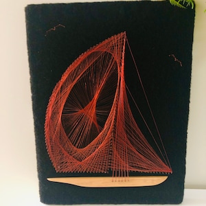 May include: String art of a sailboat on a black felt background. The sailboat is made with orange thread, with a wooden hull. Two small bird shapes are also made with thread. The artwork is displayed on a white surface.