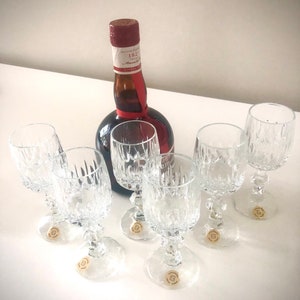 Puede incluir: Un conjunto de seis copas de vino de cristal transparente con un patr&oacute;n decorativo, junto a una botella oscura con una etiqueta roja y blanca y una cinta roja. Las copas tienen una base redonda y un tallo.