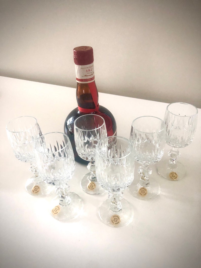 Puede incluir: Un conjunto de seis copas de vino de cristal transparente con un patr&oacute;n decorativo, junto a una botella oscura con una etiqueta roja y blanca y una cinta roja. Las copas tienen una base redonda y un tallo.