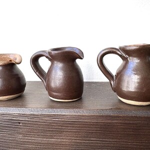 Peut inclure: Trois petits pichets en céramique marron faits à la main, avec des anses et des becs verseurs. Les pichets varient légèrement en taille et en forme, avec un bord de couleur plus claire sur le plus petit. Ils sont exposés sur une surface en bois foncé.