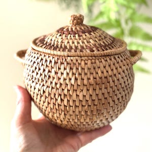 May include: A round, woven basket with a lid. The basket is made of natural-coloured fibres and features a woven pattern. The lid has a decorative knob and a darker brown pattern. The basket has two small handles on either side.