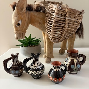 May include: A carved wooden donkey figurine with a woven basket on its back, accompanied by four small, dark-glazed ceramic pitchers featuring hand-painted floral and geometric patterns in white, blue, and orange tones on their rounded bodies.