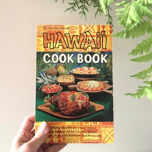 Peut inclure: Un livre de cuisine vintage intitulé "HAWAII COOK BOOK" avec une couverture jaune et orange présentant des images de plats hawaïens. La couverture comprend du texte sur les recettes traditionnelles et des photographies de plats polynésiens. Le livre est tenu dans une main.
