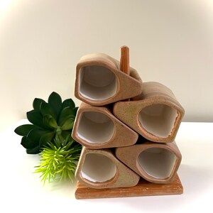 May include: A set of five ceramic mugs with a speckled tan and brown finish and white interiors, arranged in a stacked formation on a wooden rack. The mugs feature a distinct geometric, angular shape and are supported by a central wooden post.