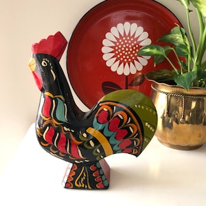 May include: A hand-painted wooden rooster figurine in black, red, green, and yellow. The folk art style rooster has a red comb and a decorative base. A red tray with a white flower design is in the background.