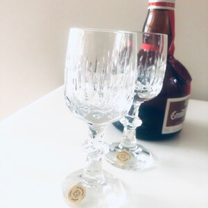 Puede incluir: Dos copas de vino de cristal transparente con l&iacute;neas verticales texturizadas, tallos y bases decorativas. Una botella de licor est&aacute; en el fondo. Cada copa tiene una peque&ntilde;a pegatina dorada en la base. Hecho en Alemania.