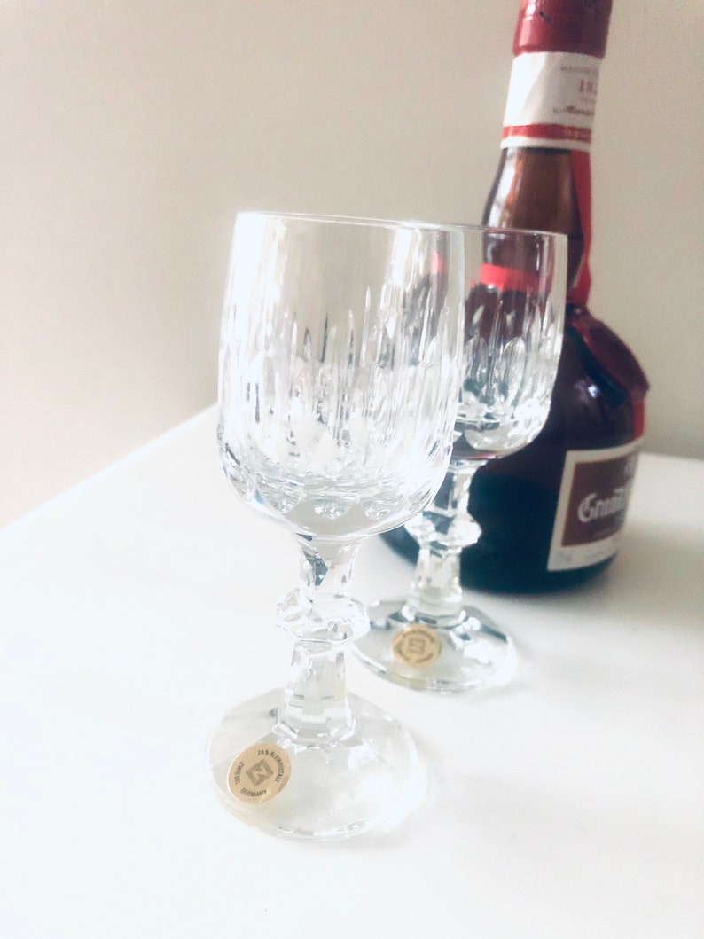 Puede incluir: Dos copas de vino de cristal transparente con l&iacute;neas verticales texturizadas, tallos y bases decorativas. Una botella de licor est&aacute; en el fondo. Cada copa tiene una peque&ntilde;a pegatina dorada en la base. Hecho en Alemania.