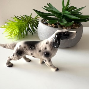 May include: A ceramic dog figurine, predominantly white with black spots and details. The dog is depicted in a standing pose, with its head turned. The figurine is on a white surface, with green and grey decorative plants in the background.