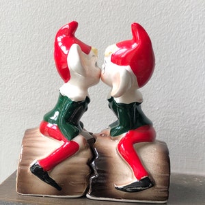 Peut inclure: Deux figurines d'elfes en céramique s'embrassant, posées sur une base en forme de bûche. Les elfes portent des chapeaux rouges, des visages blancs, des vestes vertes et des pantalons rouges. La base est marron avec une fissure au milieu.