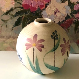 May include: A spherical, cream-coloured ceramic vase with a narrow neck. It features hand-painted purple and pink flowers, blue buds, and green leaves. The background is a floral painting.