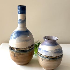 May include: Two ceramic vases with a landscape design. The taller vase has a long neck and a rounded body, while the shorter vase is round. Both feature a blue sky, beige landscape, and brown base.
