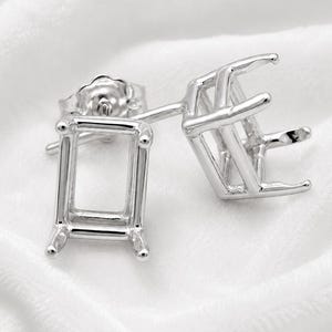May include: A pair of silver-coloured metal earring settings featuring rectangular prongs designed to hold a gemstone. Each setting includes a post and a matching butterfly clutch back, showcasing a polished metallic finish and a structural frame.