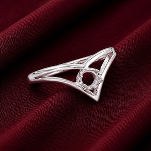 May include: A silver-colored ring with a unique open design. The ring features a V-shaped band that leads to a circular setting, ready for a gemstone. The ring is displayed on a deep red fabric.
