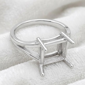May include: A silver-toned metal ring setting featuring a square-shaped basket with four prongs designed to hold a gemstone. The band has a split-shank design that tapers toward the center, providing a structural base for a custom jewelry piece.