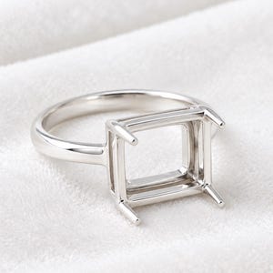 May include: A polished silver-toned metal ring setting featuring a rectangular four-prong head designed to hold a gemstone. The band is smooth and rounded, leading up to a basket-style gallery that supports the open rectangular prong setting.