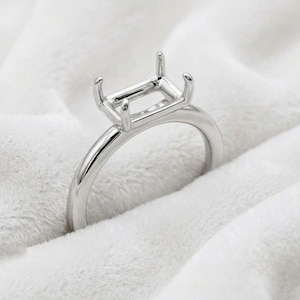 May include: A polished silver-tone metal ring setting featuring a rectangular head with four prongs designed to hold a gemstone. The band is smooth and rounded, showcasing a clean, minimalist structure for a custom jewelry piece.