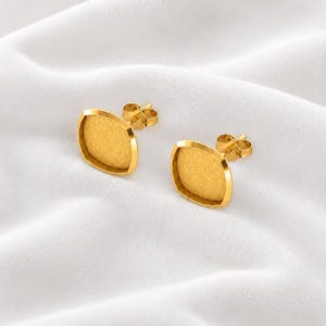 May include: A pair of gold-toned stud earrings featuring a square-shaped setting with rounded corners and a textured, matte finish. Each earring includes a standard post back mechanism for secure attachment, showcasing a clean and simple design.
