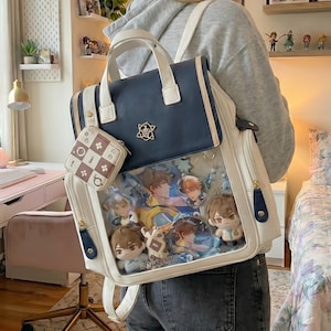 Puede incluir: Mochila blanca y azul marino con un bolsillo frontal transparente lleno de productos de personajes de estilo anime. La mochila tiene un asa superior, correas para los hombros y un pequeño monedero a juego. Un emblema dorado está en la parte delantera.