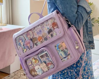 May include: A lavender ita bag with multiple clear display windows filled with anime-style character charms and cards. The bag has a top handle, a shoulder strap, and a heart-shaped zipper pull. The bag is made of a smooth, synthetic material.