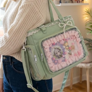 May include: A sage green tote bag featuring a clear front display window framed by pink ruffled fabric. The bag includes white lace trim along the top edge, a decorative pearl-like beaded bow, and internal anime-style character pins.