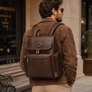 Könnte beinhalten: Ein dunkelbrauner Lederrucksack mit texturiertem Krokodilledermuster, einem Tragegriff, verstellbaren Schultergurten, einer vorderen Reißverschlusstasche sowie Seitenfächern mit silberfarbenen Metallreißverschlüssen und Beschlägen.