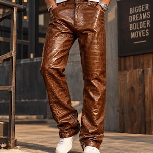 May include: Brown trousers featuring a glossy, embossed crocodile skin texture. These trousers have a classic five-pocket design with a button fly closure, a straight-leg cut, and a polished finish that highlights the intricate reptilian pattern.