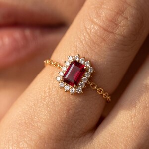 Puede incluir: Un anillo de cadena de oro con una piedra preciosa rectangular de color rojo intenso en talla esmeralda en el centro, rodeada por un halo de pequeñas piedras blancas redondas. El aro está formado por delicados eslabones dorados.
