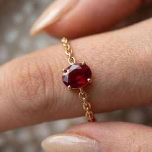 Puede incluir: Un delicado anillo de cadena de oro con una piedra preciosa de color rojo intenso de corte ovalado, engastada en cuatro garras. La banda del anillo está formada por finos eslabones de oro, creando un aspecto flexible y ligero en el dedo.