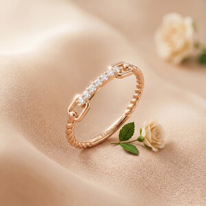 Può includere: Un anello in oro rosa con un design a fascia intrecciata e un elemento a barra orizzontale centrale incastonato con cinque piccole pietre chiare. L'anello presenta un dettaglio a maglia rettangolare su ogni lato e il marchio FERNOS inciso all'interno.